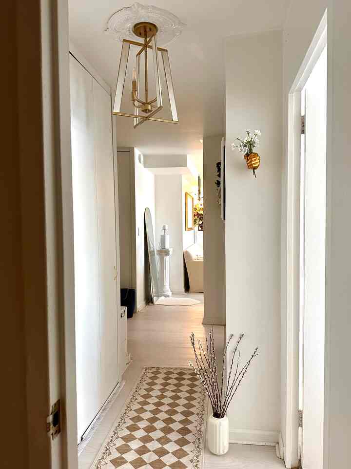 A narrow hallway in white and beige tones featuring a gold wall sconce and patterned rug creating a natural and elegant interior