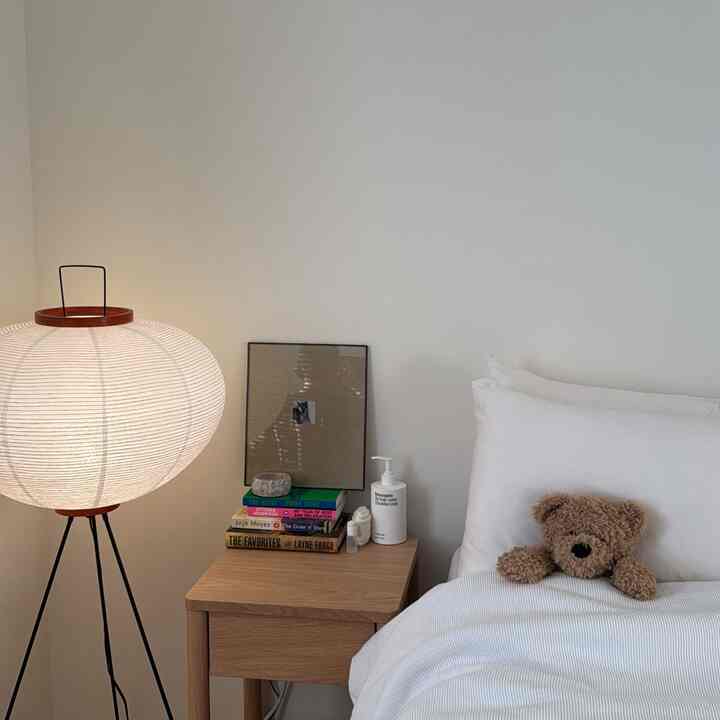 White and wood tone bedroom featuring a wooden nightstand, Japanese-style lamp, cozy bedding, and a teddy bear
