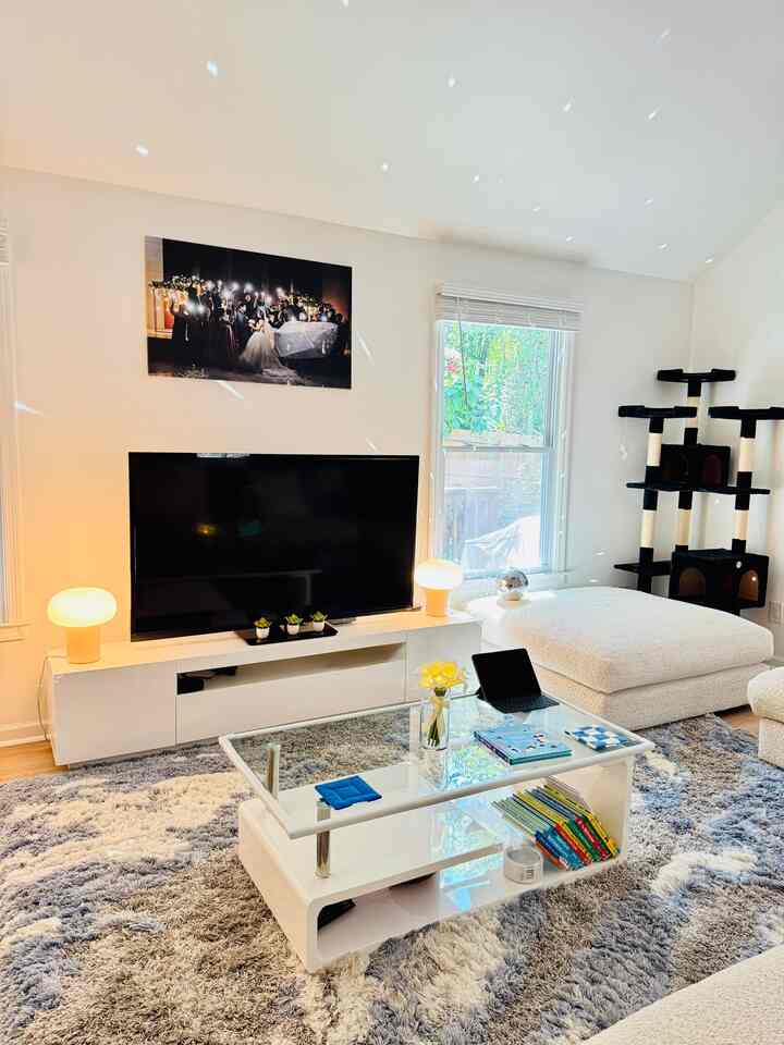 Bright white and beige toned living room featuring large TV, glass coffee table, and a cat tower in a minimal cozy space