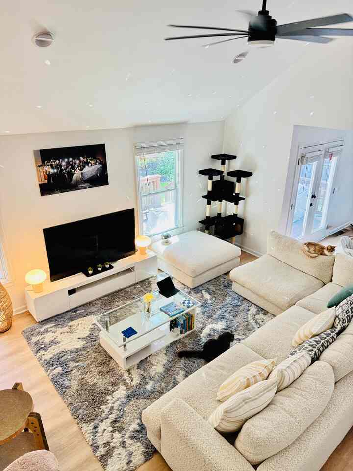 Bright natural-tone living room featuring cream sectional sofa, ottoman, a white coffee table, and a tall cat tower with two cats