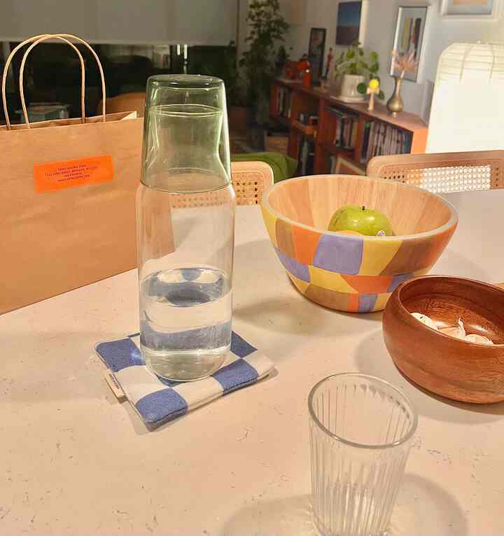 White-toned kitchen table featuring clear glass carafe on blue fabric coaster, colorful wooden bowl with green apple in a natural modern home cafe setting