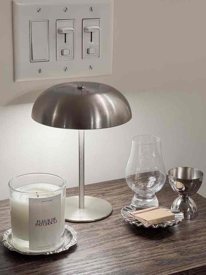 Brown wooden table featuring silver table lamp, candle, glass, and metal trays creating a neat and stylish setting
