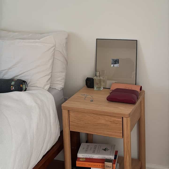 Simple bedroom with natural wood tones and white, featuring a nightstand next to the bed holding perfume and a picture frame