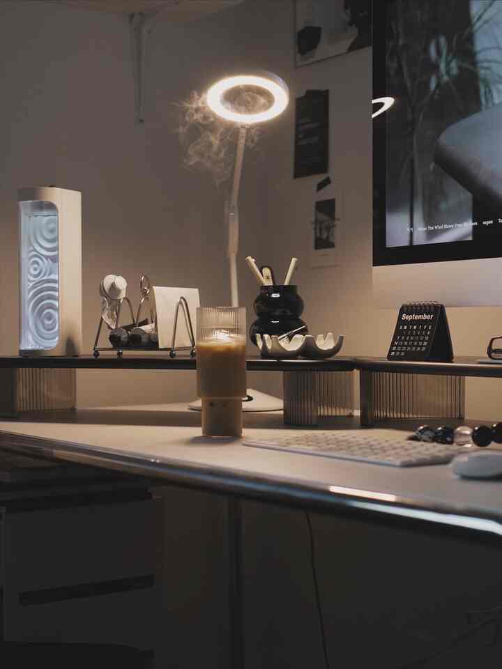 White-toned home office featuring a monitor stand, desk lamp, mini calendar, and organized workspace with a calm atmosphere