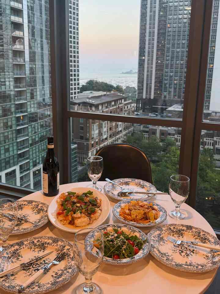 White round dining table set for four with floral plates and wine glasses near large window showing urban cityscape at sunset for home party