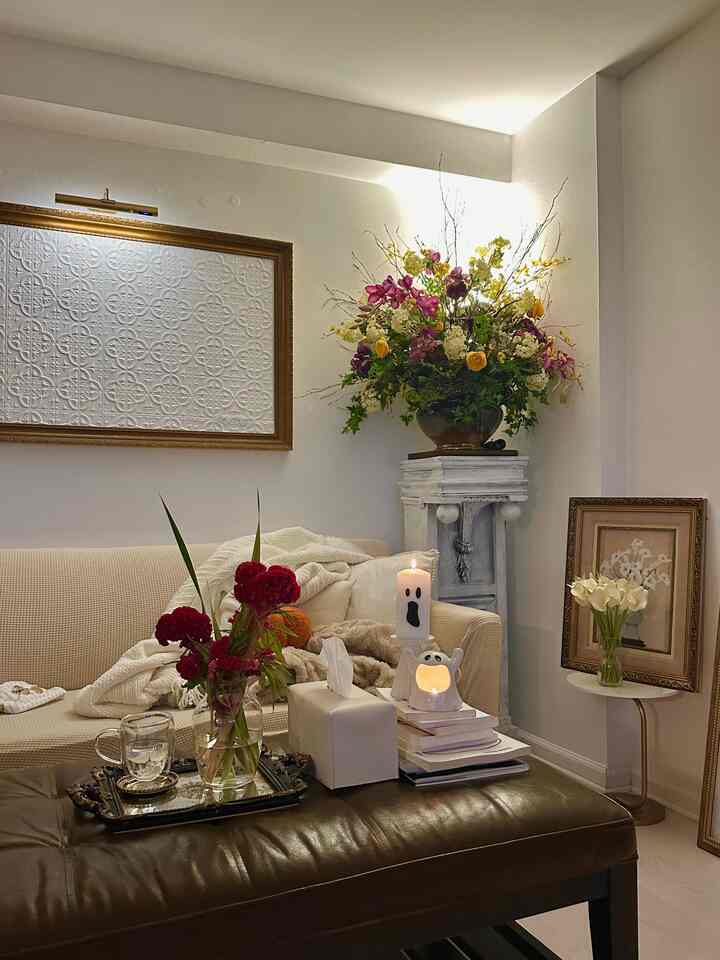White and beige toned living room featuring abundant floral vase on pedestal and dark brown leather ottoman, creating a romantic French country atmosphere