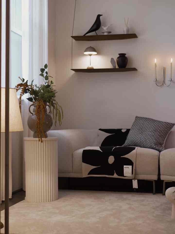 Beige and white toned living room featuring a sofa, side table with vase, and shelves with lighting on the wall creating a cozy atmosphere