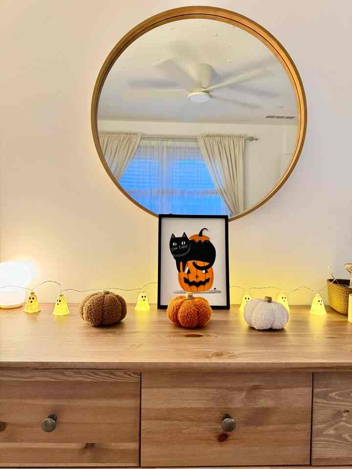 Beige-walled baby room with round wooden mirror, featuring Halloween-themed black cat print, ghost string lights garland, and plush pumpkins on wooden dresser, creating cozy vibe