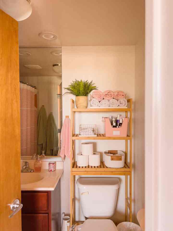 Small bathroom corner with soft pink and wood tones, neatly organized towels and a plant creating a natural, tidy atmosphere