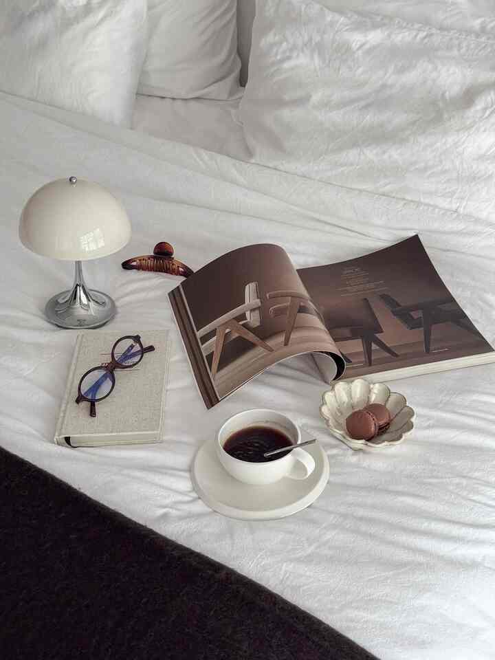 Bright white-toned bedroom featuring a minimal table lamp, coffee cup, and a cozy setup with book and glasses
