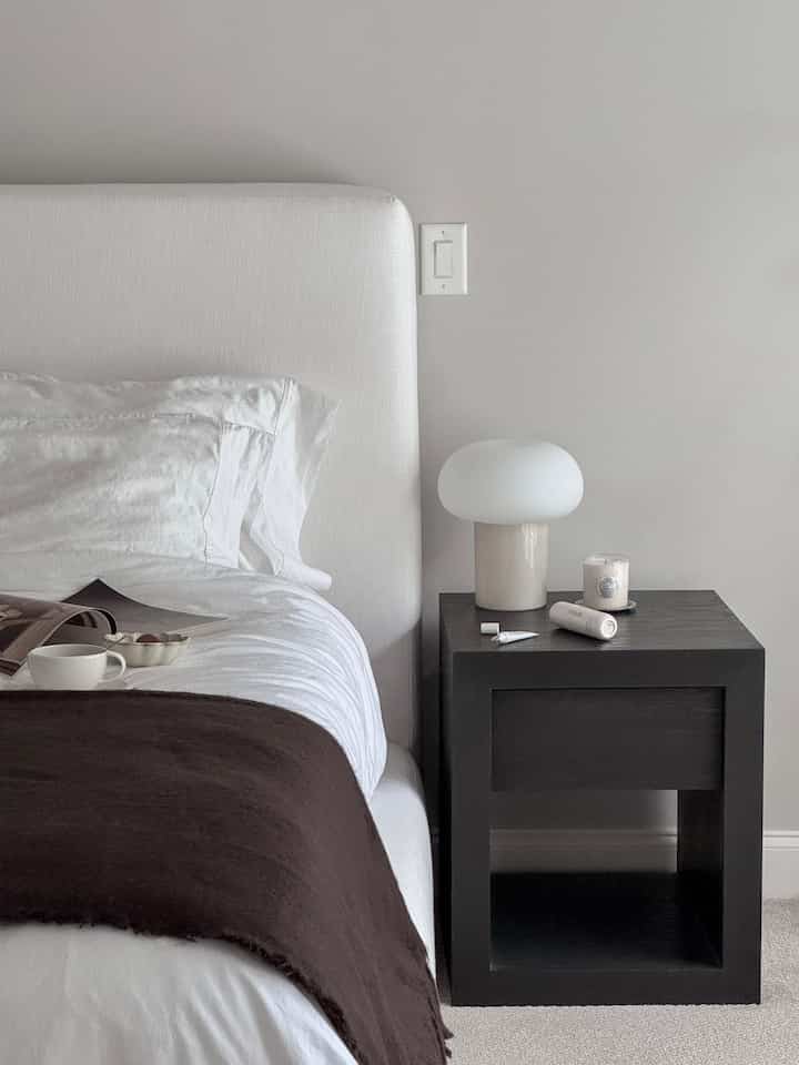 White and dark brown toned bedroom featuring a white bed with dark brown throw and black nightstand topped with a table lamp, creating a cozy atmosphere