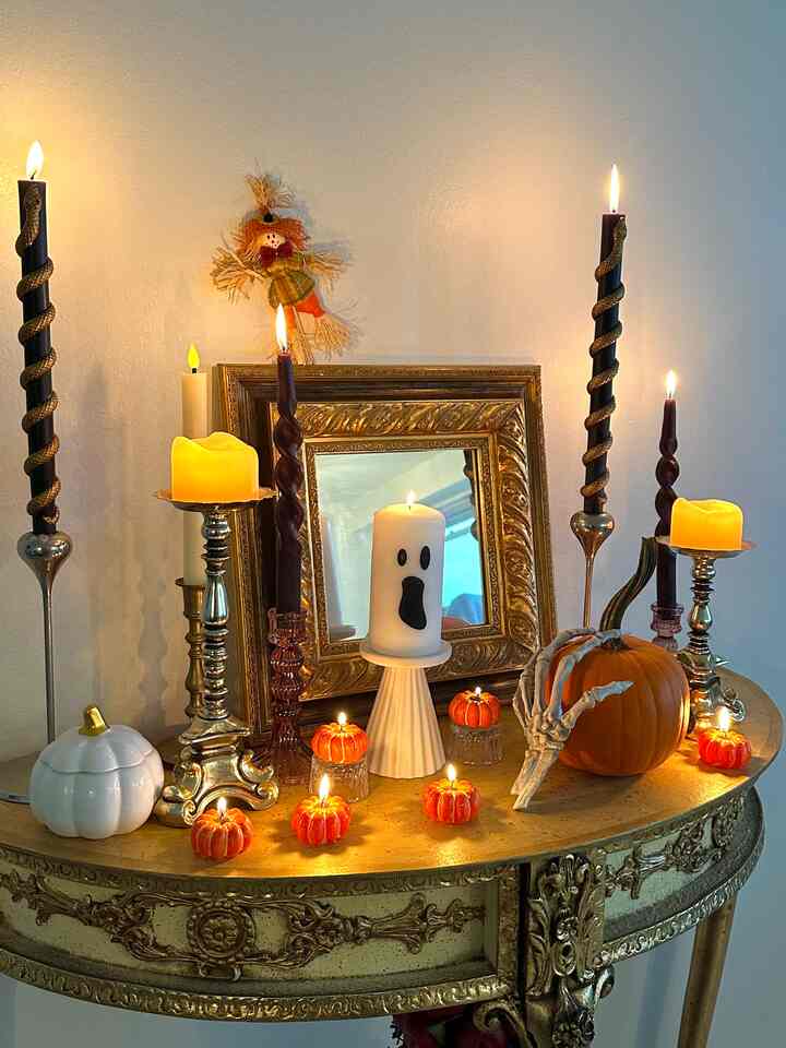 White wall and ornate gold table featuring black candles, orange pumpkins, and a skeleton hand figurine in a Halloween decor setup