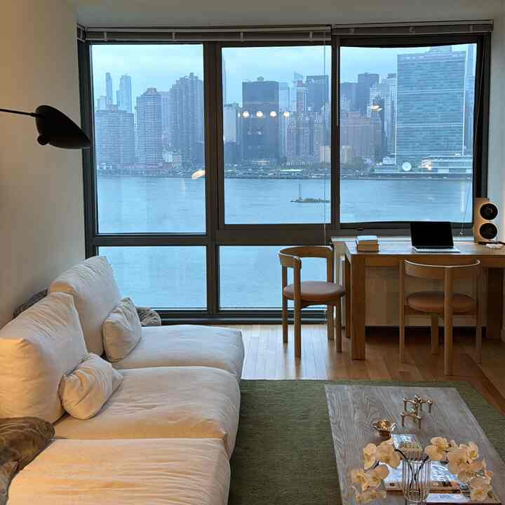White sofa and natural wooden desk in a Japandi-style living room featuring large windows overlooking a city river, creating a cozy atmosphere