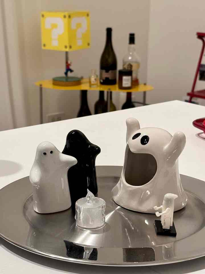 White and black ghost-shaped ceramic ornaments on a silver tray atop white table, with yellow question-block lamp and bottles in background