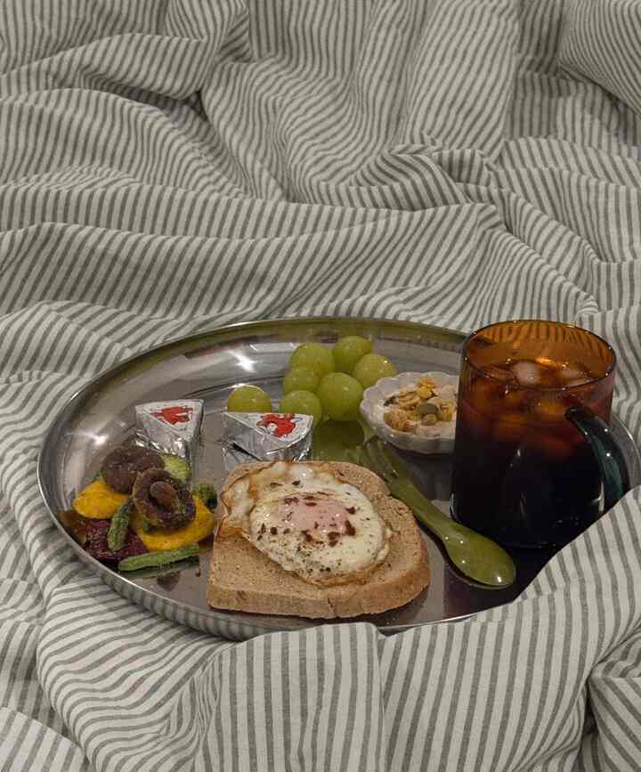 Cozy bedroom scene with striped duvet cover and silver tray holding toast with egg, fruits, and a cold drink for breakfast