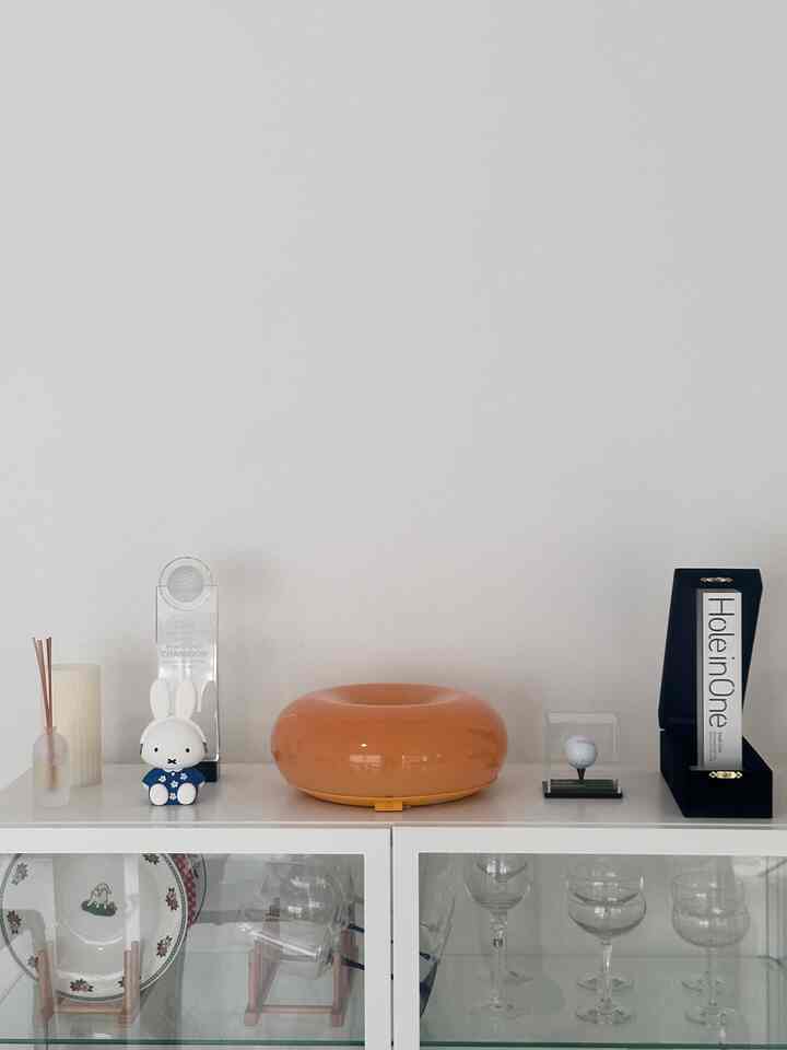 Minimal white dresser space topped with an orange donut lamp and neat decorative objects against a white background