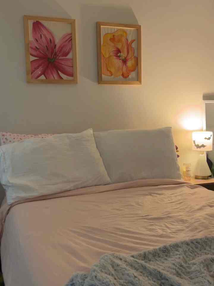 Cozy bedroom with pink bedding, white pillows, warm table lamp lighting, and floral framed artworks on the wall