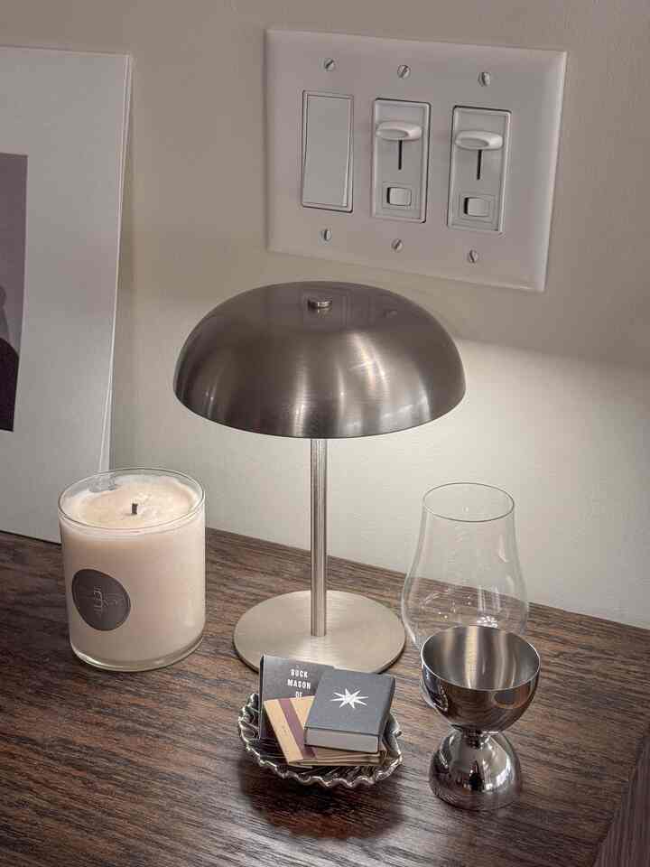 A minimal home bar setting featuring a silver table lamp, candle, and metallic bowl on a wood tone surface with ambient lighting