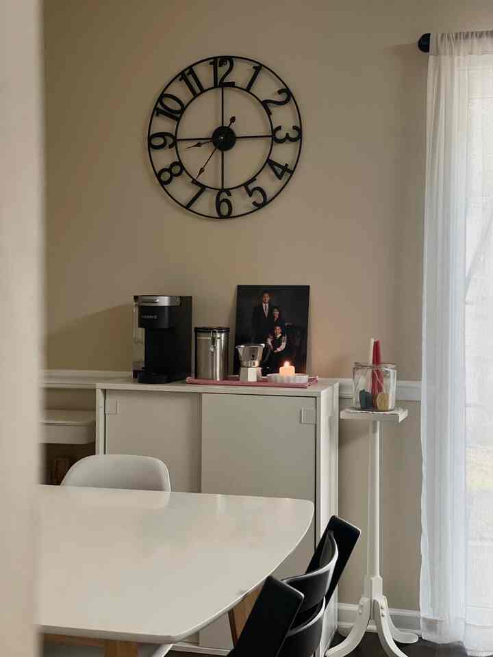 Soft beige walled dining room featuring white furniture, a large black wall clock centered above cabinets, a coffee maker, and a lit candle creating a cozy atmosphere