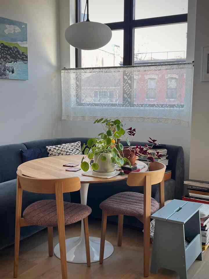 A cozy dining room with a blue velvet corner sofa, round wooden dining table, dining chairs, and plants arranged near window with pendent lighting