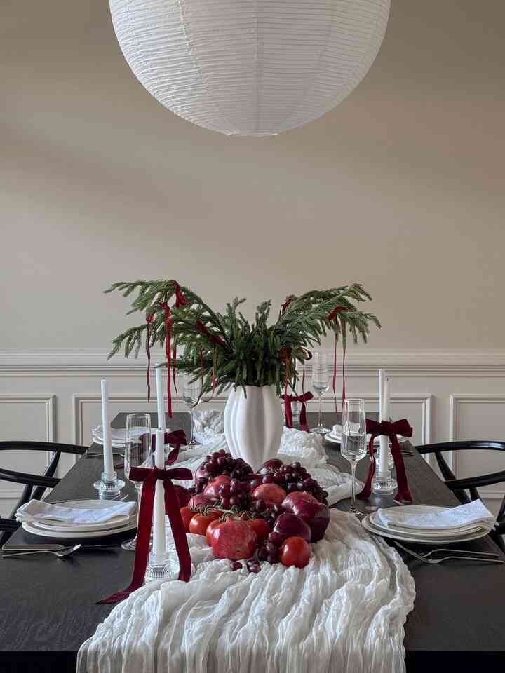 Beige-walled dining room with white pendant light, featuring dark wood dining table decorated with white vase of green foliage and candles tied with red ribbons