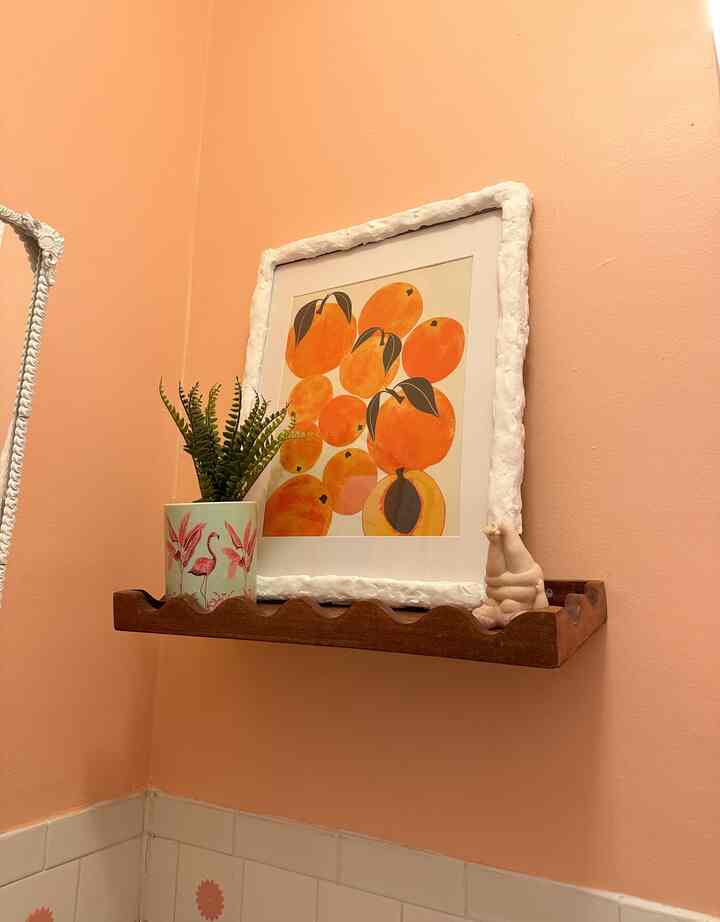 Pastel peach-walled bathroom corner featuring a wooden wavy shelf decorated with peach art print and a pink flamingo plant pot