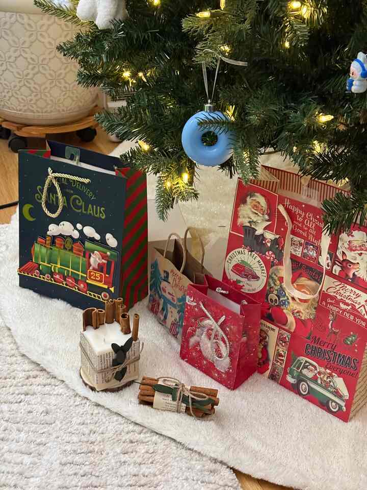 A cozy living room scene with white and beige tones featuring a Christmas tree, gift wrapping bags, and cinnamon candle decor