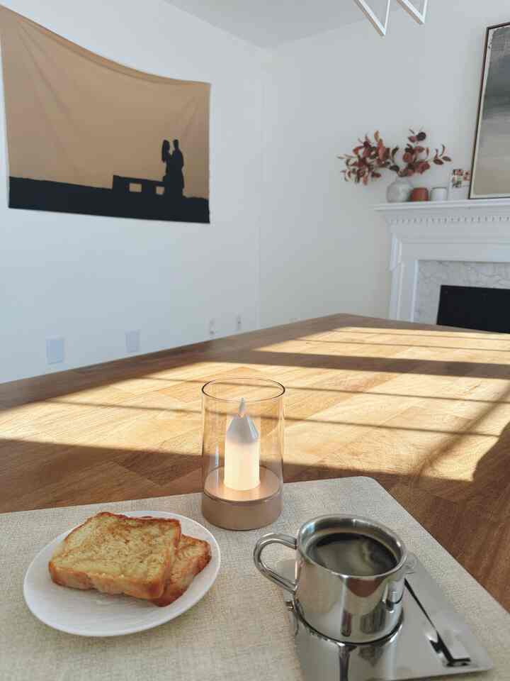 Simple dining room with white walls and wood tone table, featuring coffee and toast beside a table lamp