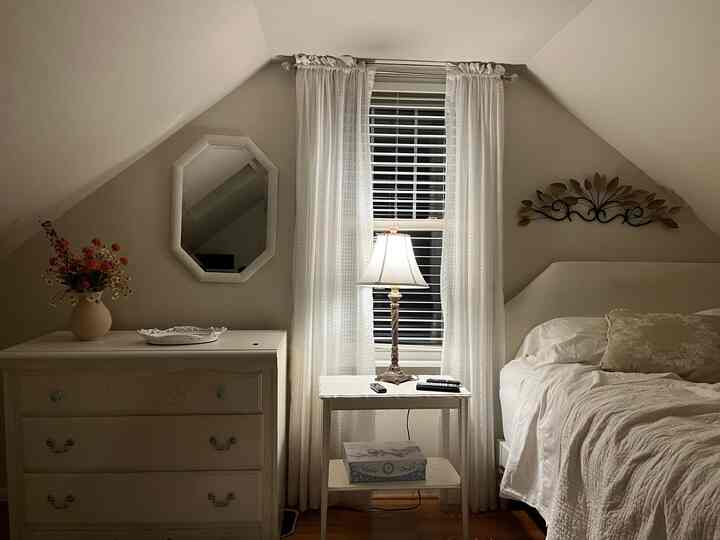 An ivory and white toned bedroom featuring a slanted ceiling, white bed, white curtains, and a nightstand table lamp creating a cozy atmosphere