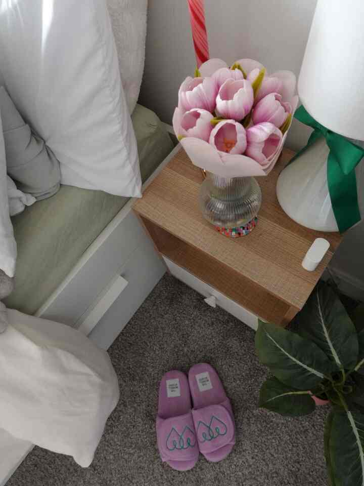 Cozy bedroom corner featuring white bedding, purple accents on slippers and tulip bouquet, natural wood nightstand, and green plant