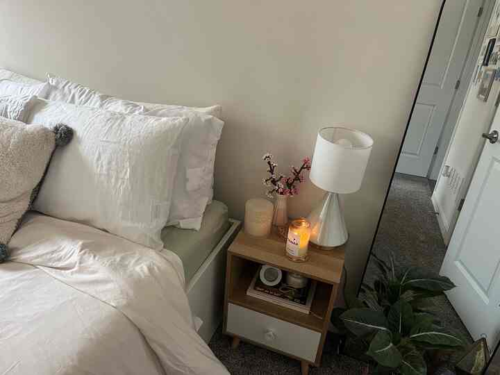 A clean bedroom with white and beige tones, featuring a nightstand with a lamp and candle