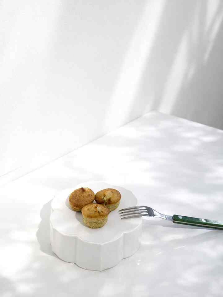 Minimal white-toned setting featuring a white plate with three muffins and a green-handled fork on a bright surface