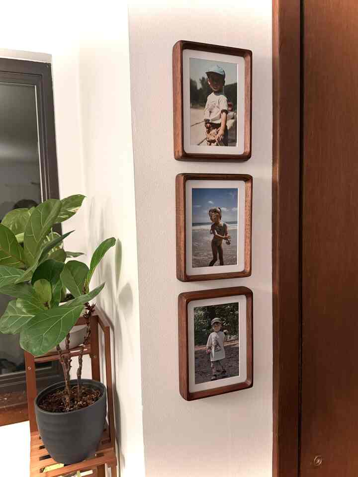 Beige-toned wall corner featuring three vertically aligned wooden picture frames with a potted plant on a wooden stand beside it.