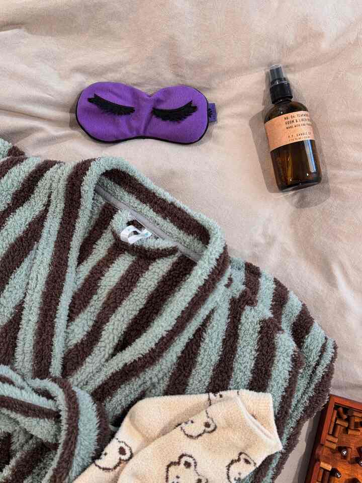 Beige-toned bedding with a purple sleep mask, striped brown bathrobe, and a room spray bottle arranged for a cozy feel