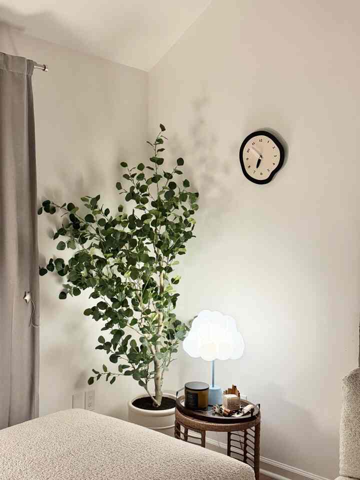 Soft white-toned living room corner featuring a large potted plant, round wicker side table with cloud-shaped table lamp, and a wall clock, creating a warm and simple ambiance