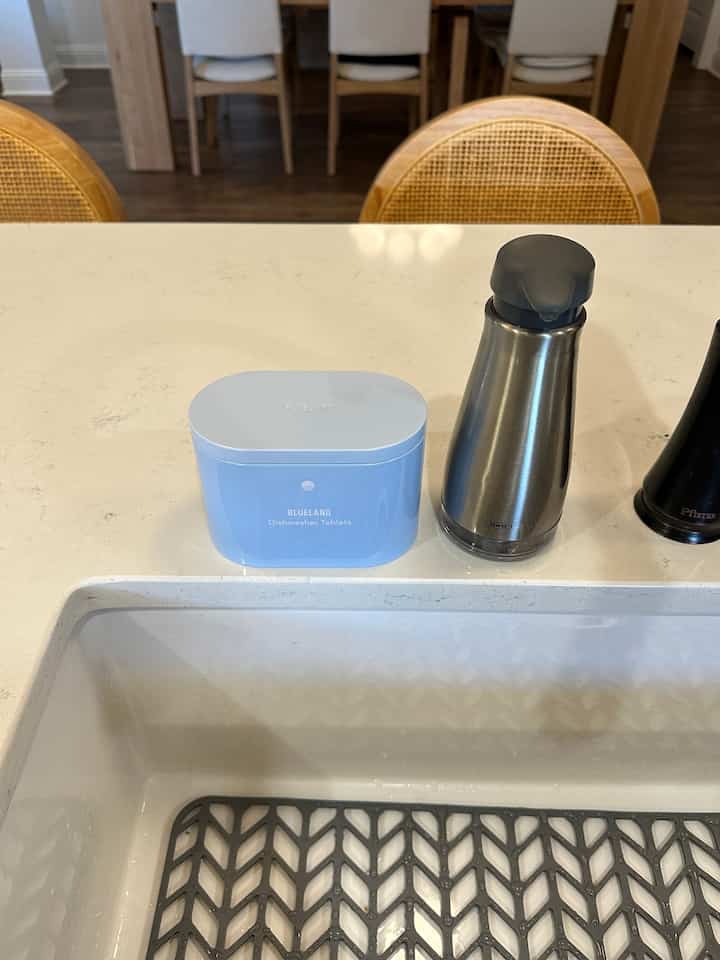 A natural-tone and white kitchen featuring a blue dishwasher detergent container and stainless steel soap dispenser on the countertop
