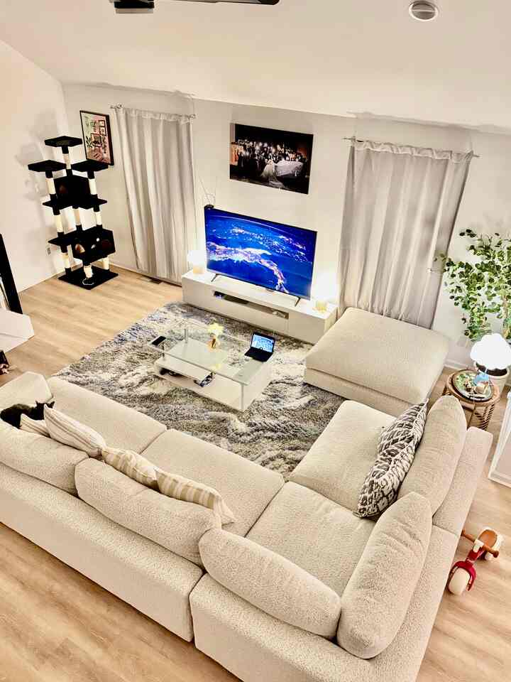 Bright beige natural modern living room featuring an L-shaped sectional sofa, glass coffee table, cat tower, and warm lighting for a cozy atmosphere