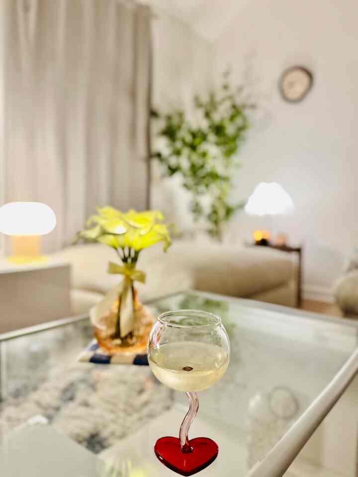 A beige and natural tone living room featuring a glass coffee table with a heart-shaped wine glass and vase, creating a cozy atmosphere