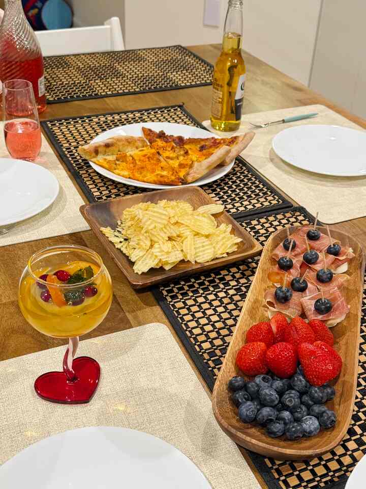 Cozy kitchen dining table with wood tone surface featuring pizza, chips, strawberries, blueberries, and a red heart-shaped wine glass for a home party setting