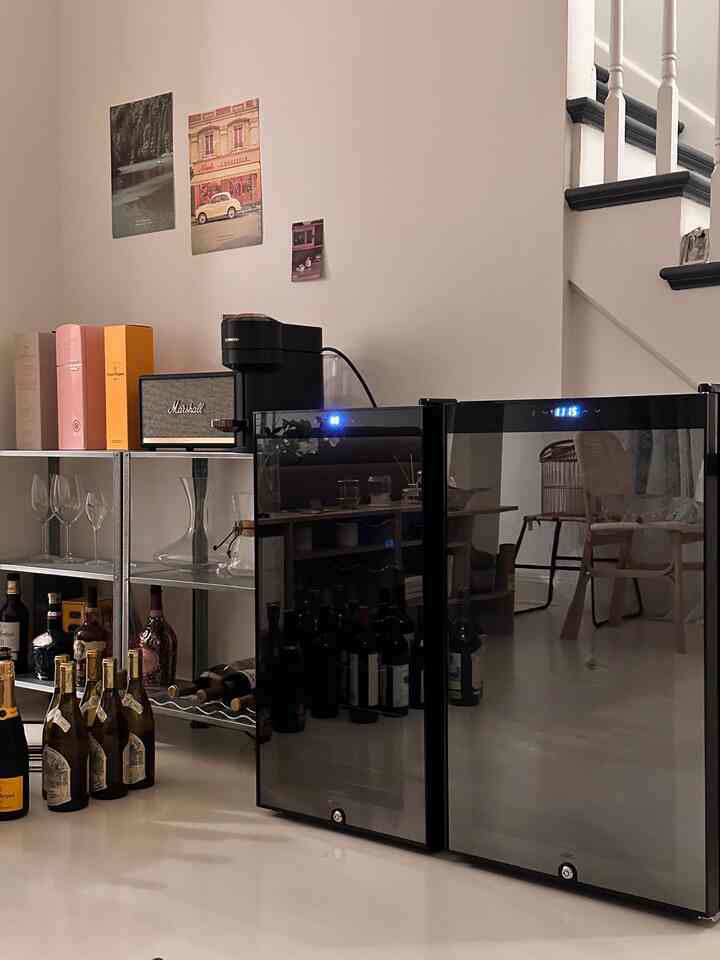 Modern kitchen space with white walls and floor, featuring black wine refrigerators and metal shelving holding a coffee machine and wine bottles, creating a sleek home cafe atmosphere