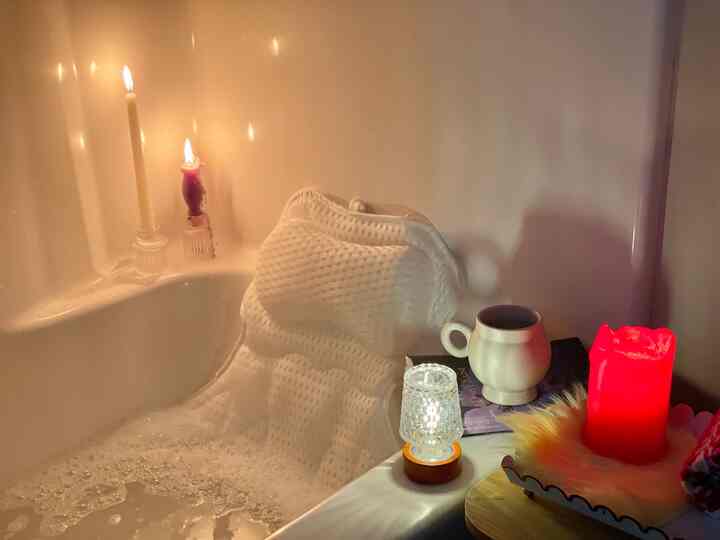 White-toned bathroom featuring a bathtub with foam, lit candles, and a beige mug creating a cozy relaxing atmosphere