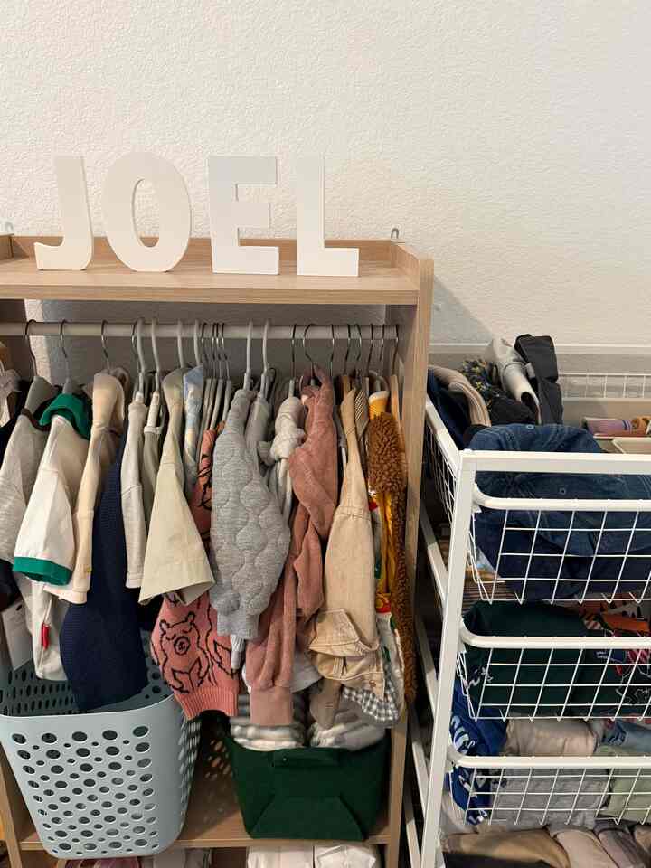 White-walled studio apartment with wood tone wardrobe, clothes hanging, and storage baskets in a compact space