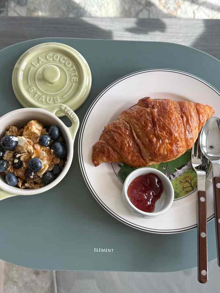 Brunch set on pastel green tabletop featuring croissant and blueberry granola with brown-handled cutlery