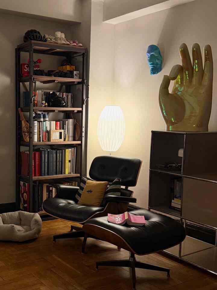 Warmly lit modern living room reading corner featuring an Eames Lounge Chair, ottoman, pet bed and bookshelf with cozy atmosphere