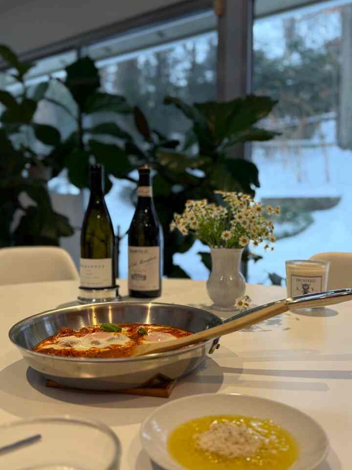 White dining room with a central stainless steel frying pan, wine bottles, and vase, creating a natural atmosphere
