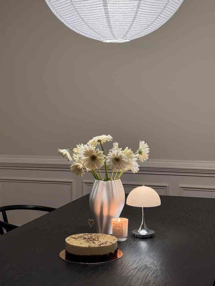 White and black toned dining room featuring a central vase, table lamp, cake, and candle creating a cozy simple modern atmosphere