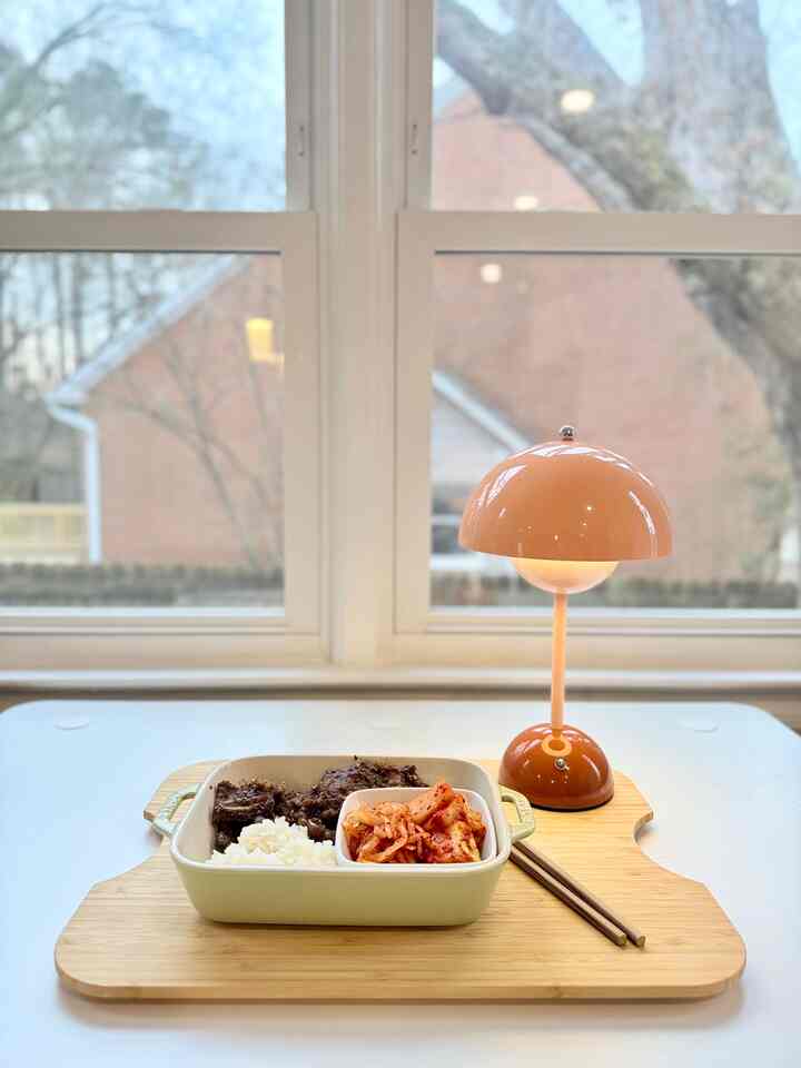 White table featuring a warm brown mushroom-shaped wireless charging table lamp and stoneware dish with LA galbi and kimchi in a dining area by a window