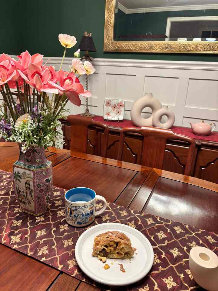 Cozy vintage dining room with green walls, wooden dining table adorned with vase of flowers, coffee mug, and apple dessert
