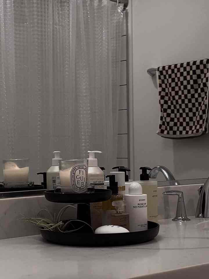 Gray-toned bathroom countertop with a black tray holding various cosmetics and candles, and a checkered towel hanging on the wall, presenting a simple bathroom space