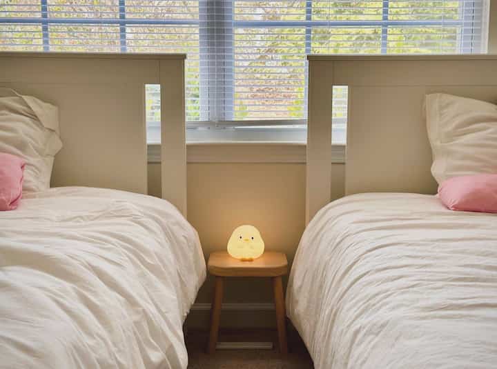 White-toned twin kids' bedroom featuring two beds with a wooden stool holding a cute chick-shaped night light, creating a cozy atmosphere